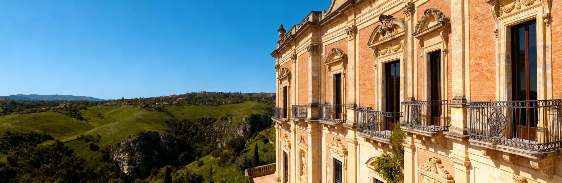 Dimora storica a Ragusa, con facciata barocca in mattoni, circondata da colline verdi e cielo azzurro.