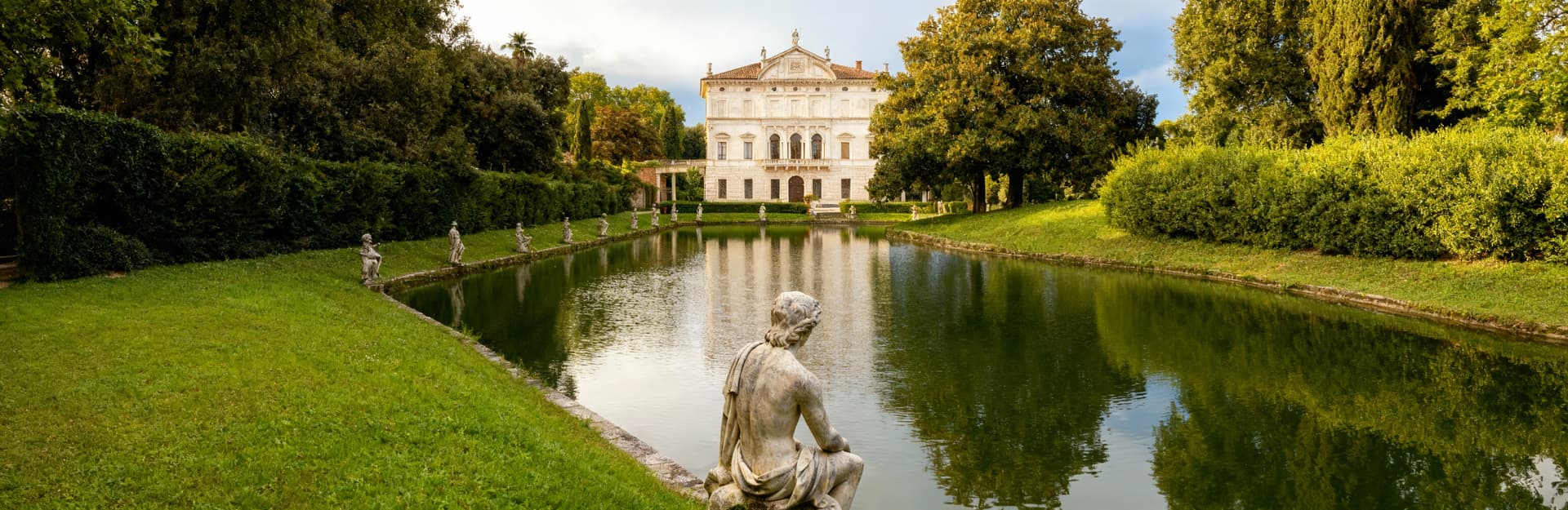 Villa storica a Mantova con facciata in pietra chiara, giardini lussureggianti e lago sereno in un contesto di lusso.