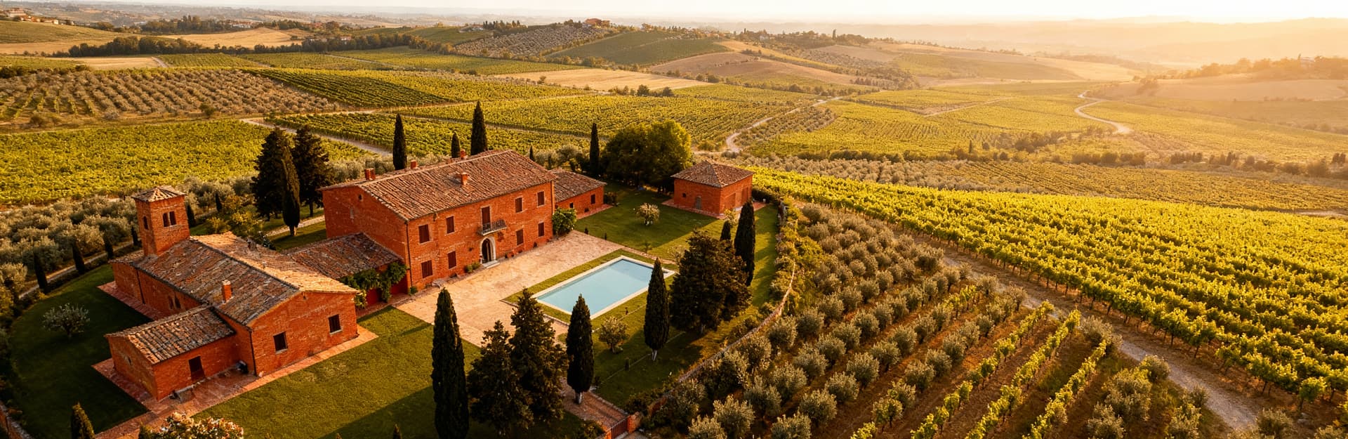 Vista aerea di un'azienda agricola toscana con uliveti e vigneti, strutture in mattoni rossi incastonate nel paesaggio.