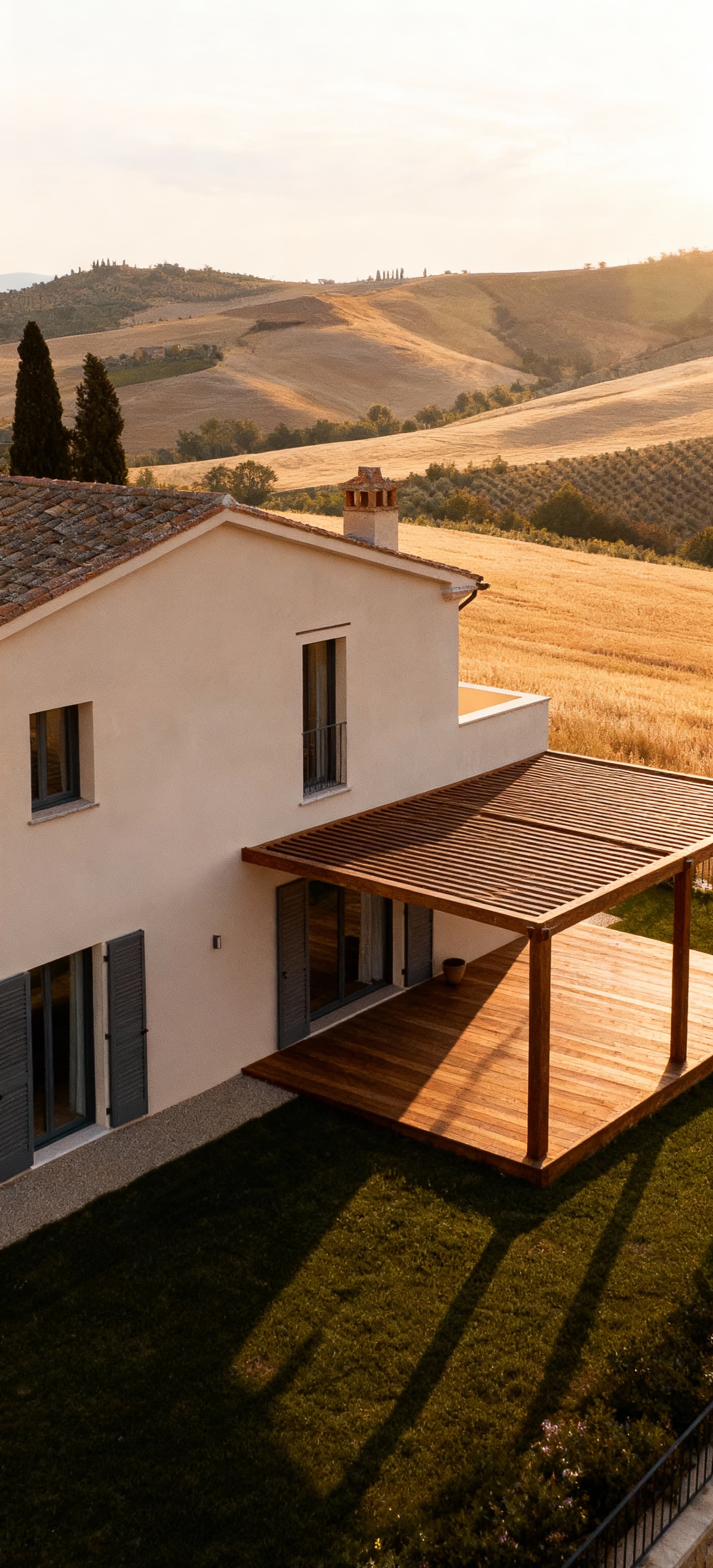 Villa esclusiva in Val d'Orcia, circondata da colline e campi dorati, con facciate chiare e terrazze in legno, nella luce del pomeriggio.